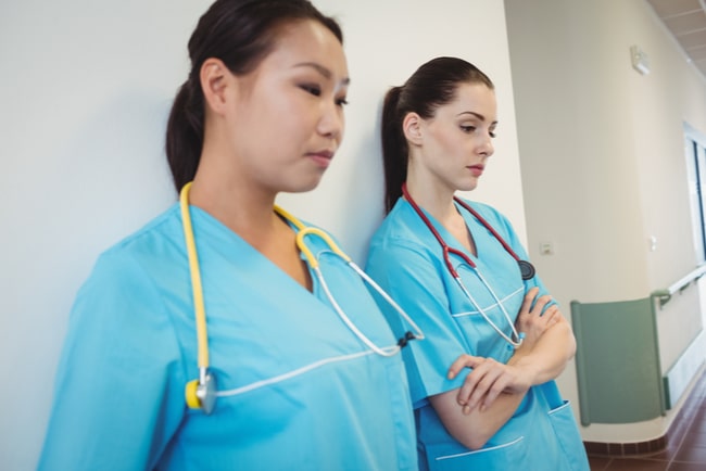 Två sjuksköterskor står lutade mot en vit vägg och ser trötta ut.